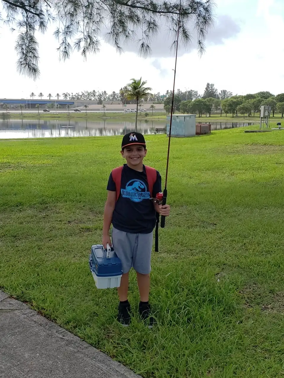 A young boy in a Jurassic World shirt holding a fishing rod and tackle box beside a suburban lake