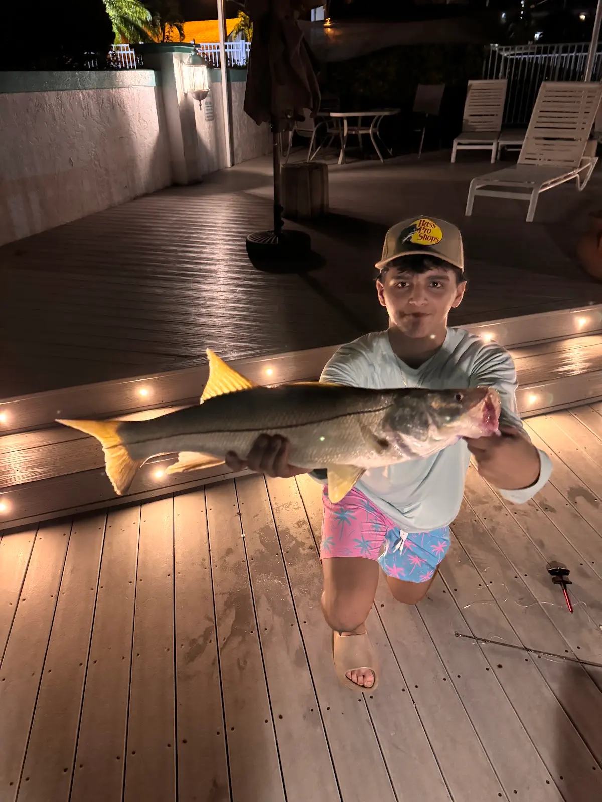 A teenager kneeling on a lit dock at night holding a large snook