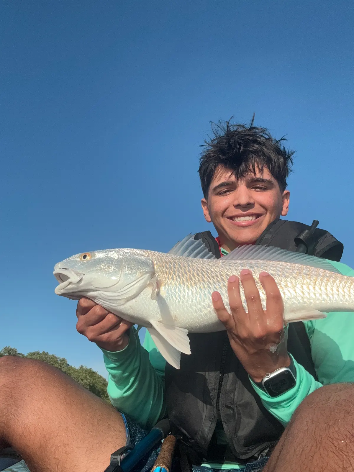 A teenager smiling and holding up a redfish against a clear blue sky