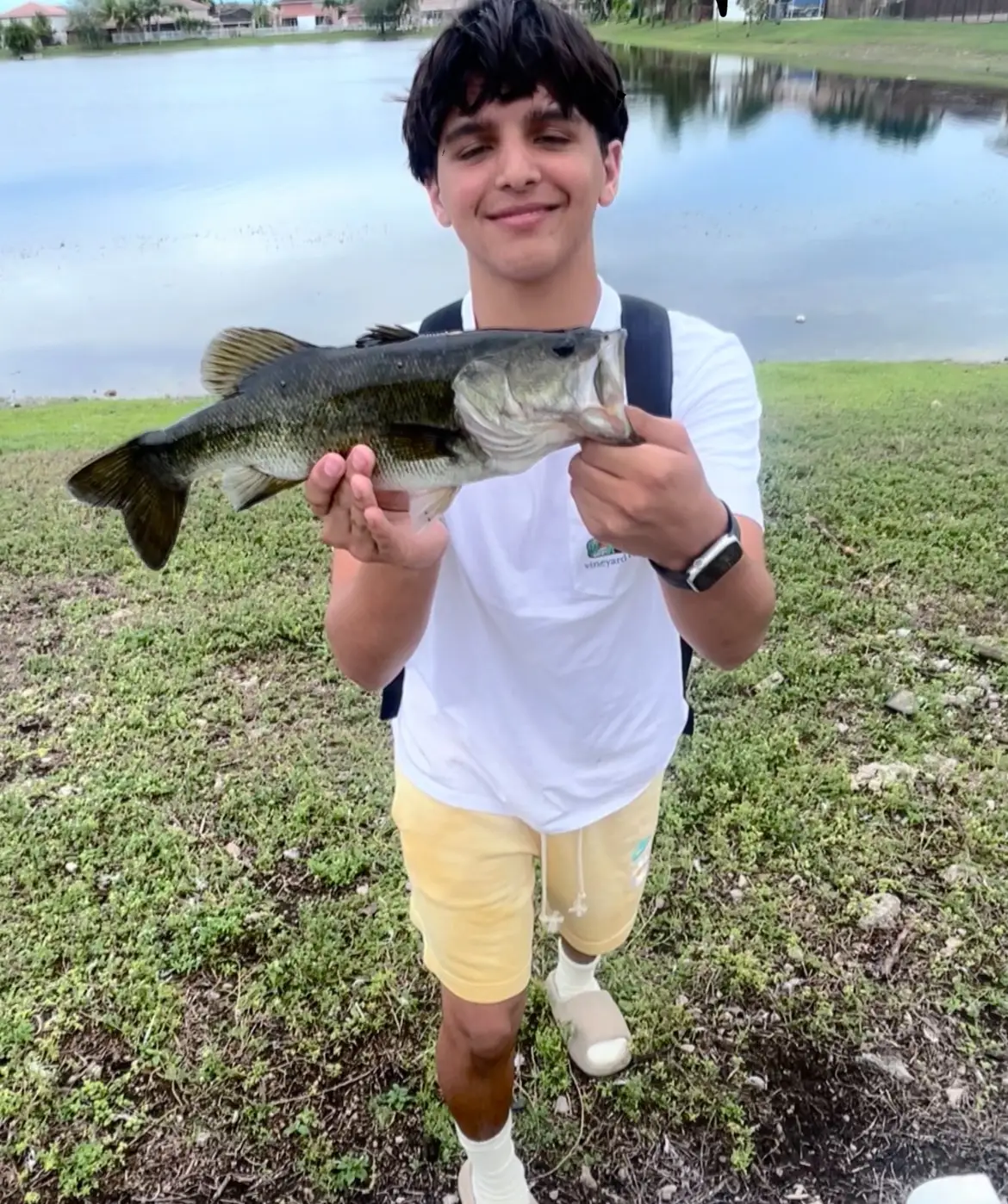 A teenager holding a largemouth bass by a neighborhood lake, backpack still on