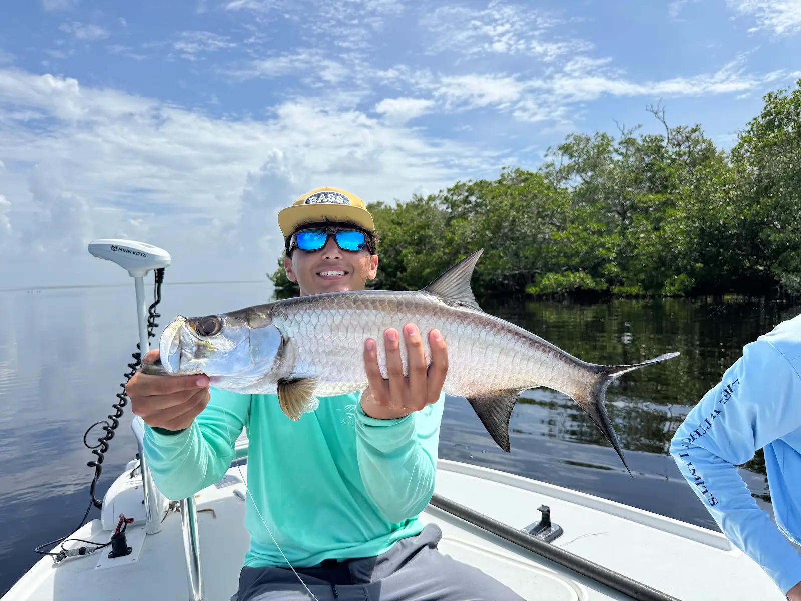 A teenager in an Anglers Club shirt holding a tarpon on a flats boat near mangroves
