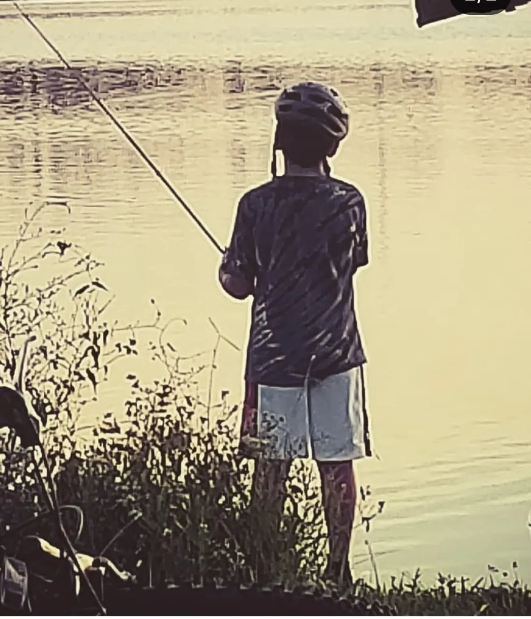 A boy with his bike helmet still on, fishing at the water's edge at golden hour