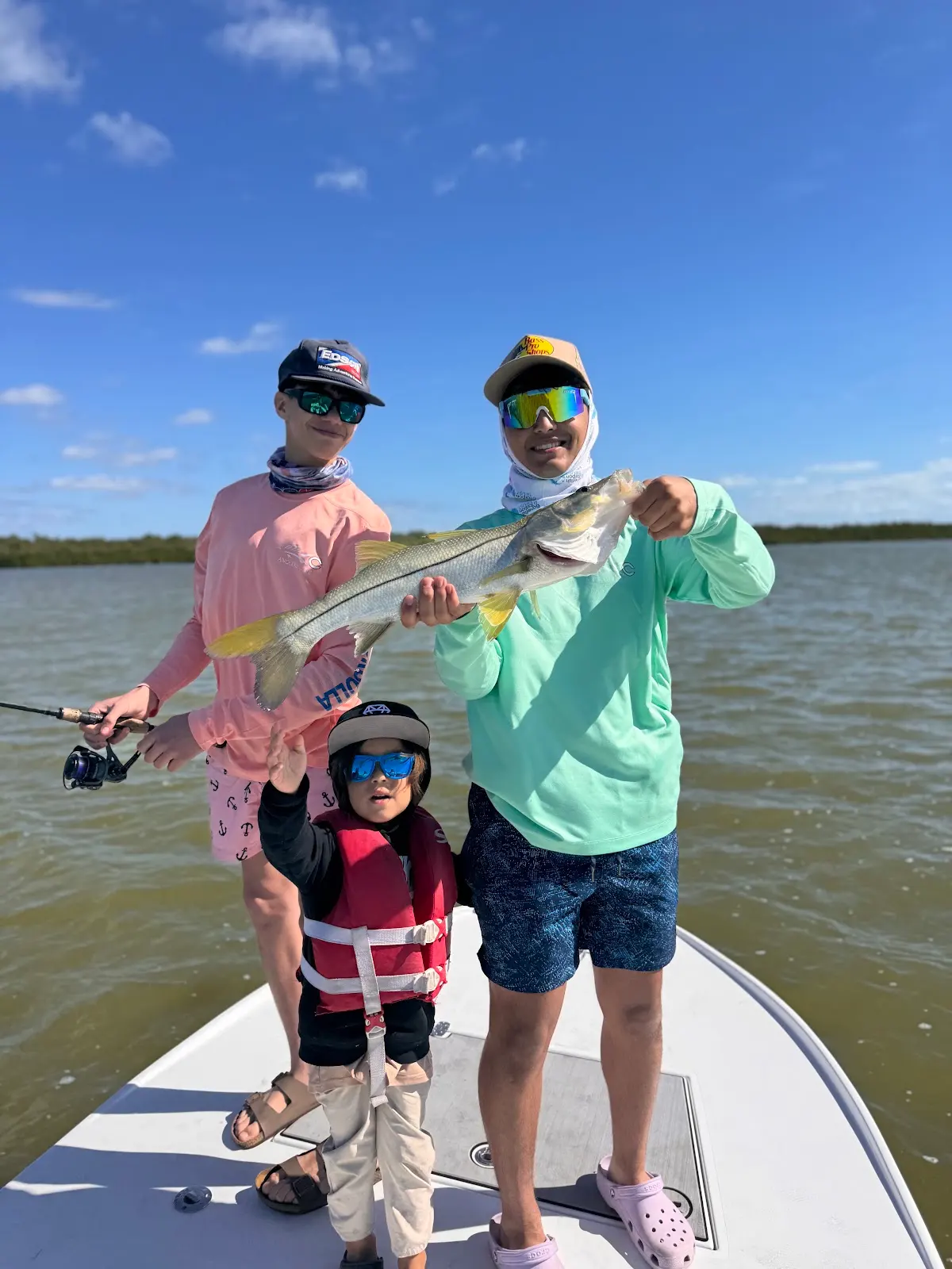 Two teenagers and a younger kid on a boat bow, holding up a snook