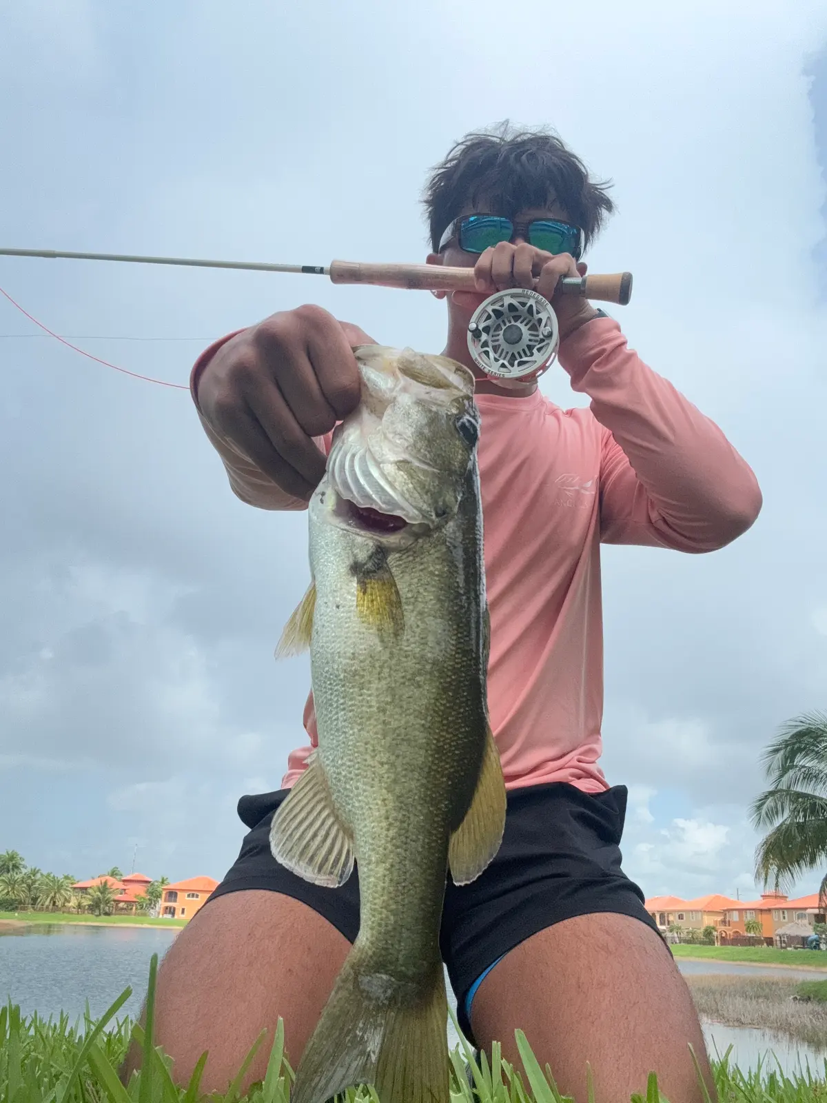A teenager holding a fly rod and reel up to his face, largemouth bass in his other hand