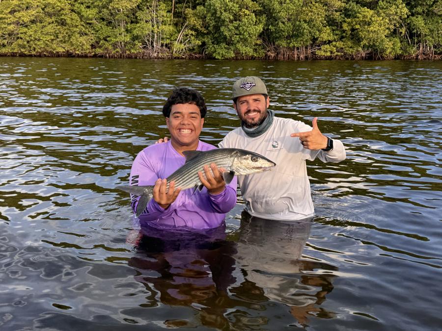 A teenager in a purple shirt and a bearded man in white, standing waist-deep in water holding a bonefish, mangroves behind