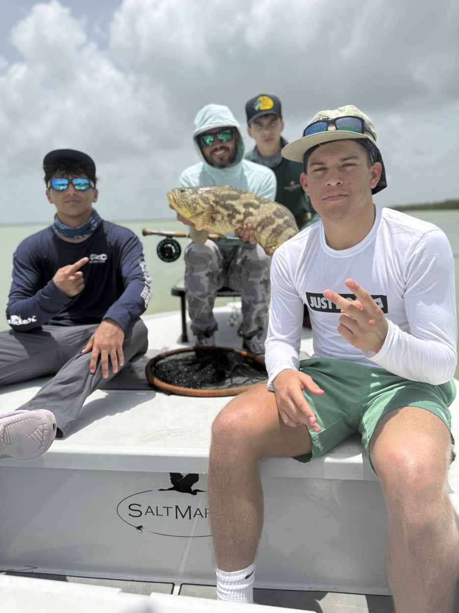 A bearded man holding a large grouper, flanked by three teenagers on a fishing boat